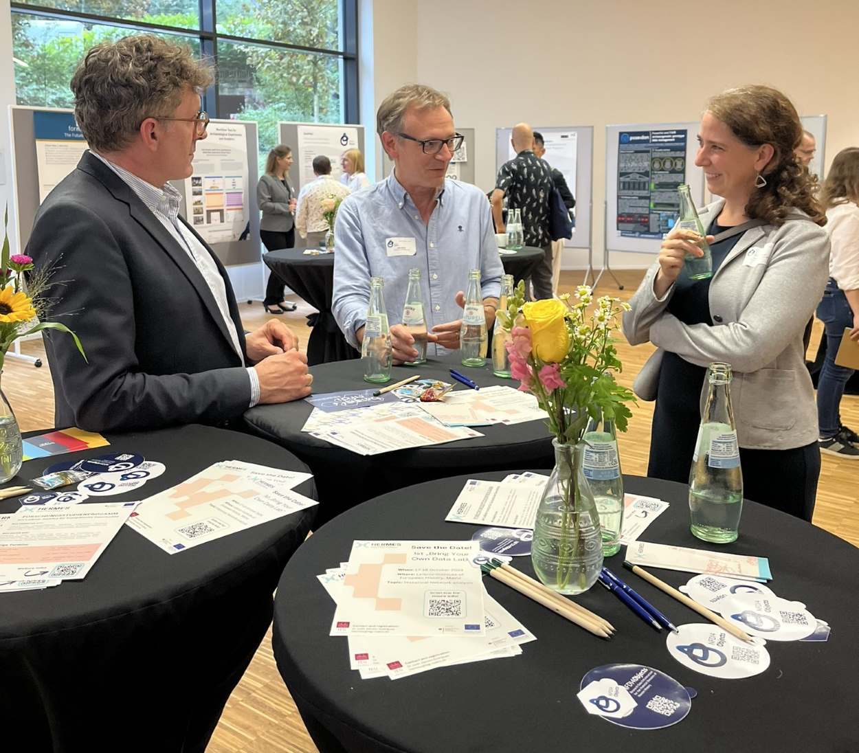 Zwei Männer und eine Frau stehen um einen Stehtisch neben Posterstellwänden und unterhalten sich.