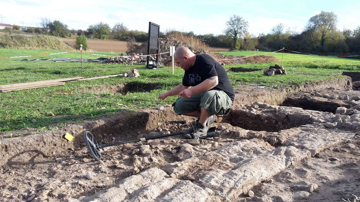 Ein Sondengänger mit Nachforschungsgenehmigung im Einsatz auf einer Grabung im spätantiken Burgus von Eisenberg Die Grabung ist ein Kooperationsprojekt der Generaldirektion Kulturelles Erbe Rheinland-Pfalz mit dem Institut für Klassische Archäologie der Universität Heidelberg. Die Einbindung von Interessierten aus der Öffentlichkeit wie Sondengänger:innen ist uns genauso wichtig wie die Einbindung der Fachcommunities in NFDI4Objects. Eine Ausgrabungsstätte ist zu sehen. Ein Mann kniet auf dem Boden. Neben ihm liegt ein Metalldetektor. Der Mann legt etwas nicht erkennbares in eine Box neben der Ausgrabungsstätte.