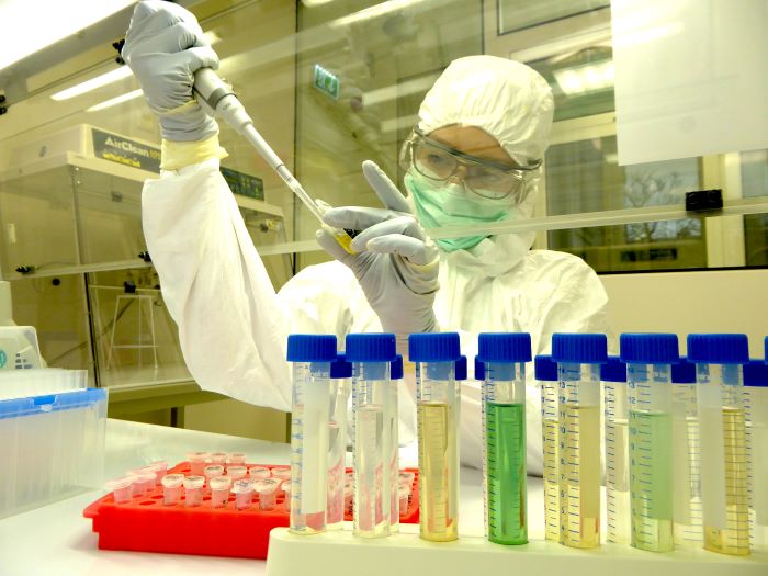 Labormitarbeiterin im Reinraum bei der Arbeit. Eine Person in weißer Schutzkleidung, einer Schutzbrille und Maske nimmt mit einer Pipette eine Probe in einem Labor.